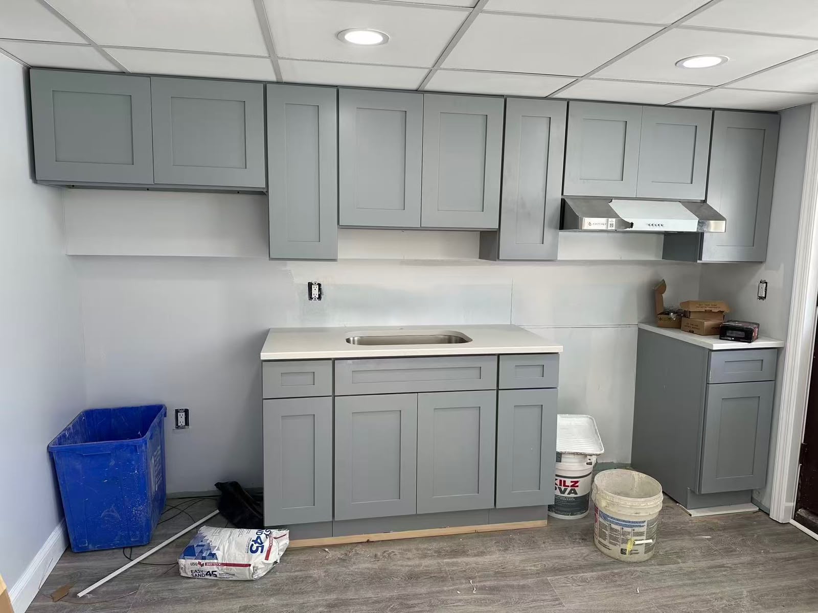 Grey shaker cabinets and counter fitted in a drop ceiling room, compound bucket still on the floor