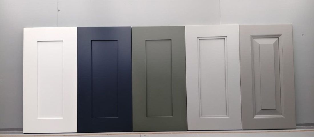 Shaker cabinet door samples in white, navy, sage and light grey, with a warm grey raised panel door, leaning against a wall